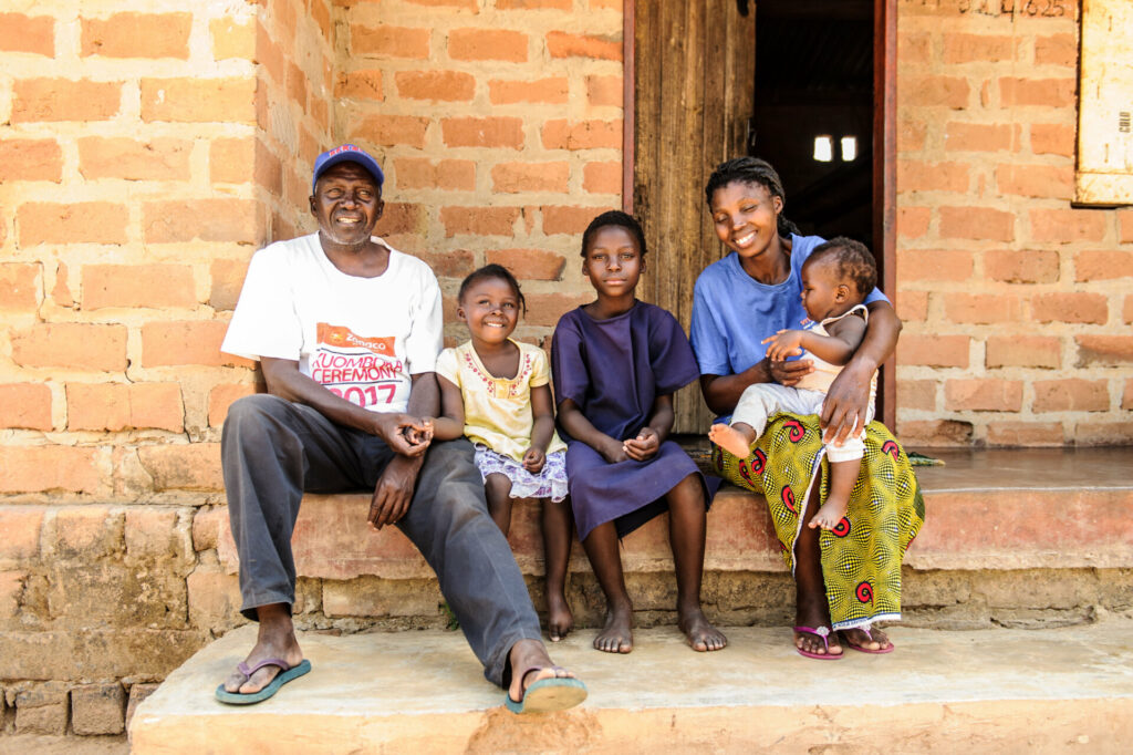 Photo: Family sitting in front of their home, which is lit up at night by an off-grid connection through a Solar Home System solution – Jason Mulikita for BGFA.